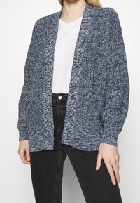Stickad cardigan med ett fläckigt blått mönster, öppen fram, långa ärmar, textil med struktur och avslappnad passform, matchad med en vit skjorta och jeans.