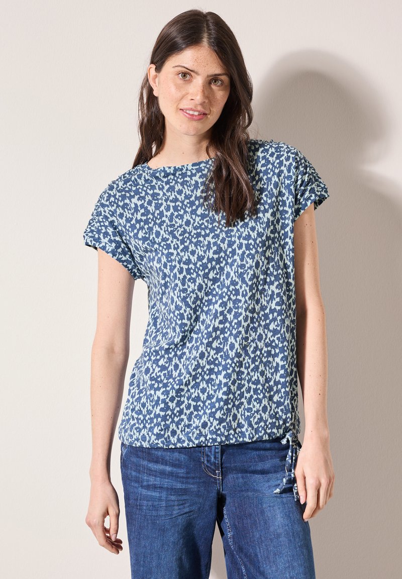 Camicia a maniche corte con stampa floreale blu e scollatura a girocollo, con lacci all'orlo. Il tessuto ha una consistenza morbida. Indossata con jeans blu.