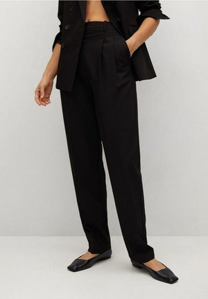 Woman wearing high-waisted black tailored pants with side pockets and black pointed flats, paired with a black blazer with rolled sleeves.