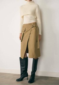 Jupe beige évasée avec un lien latéral et des poches, associée à un pull blanc côtelé court et des bottes en cuir noires jusqu'aux genoux.