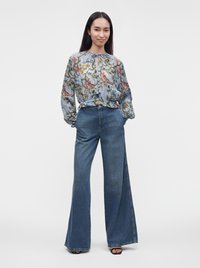 Femme portant un chemisier bleu clair à motif floral à manches longues et un jean bleu taille haute à jambes larges, debout les mains dans les poches.