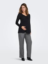Haut noir à manches longues avec un col en dentelle, associé à un pantalon gris ajusté. Le modèle montre un ventre arrondi, mettant en avant le confort des vêtements de maternité.