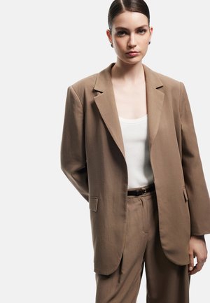 Blazer marrón a medida en una tela texturada, con cuello de solapa, un botón y bolsillos frontales. Se combina con una camiseta sin mangas blanca de canalé.