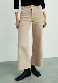 Jeans beige a gamba larga con orli sfrangiati, realizzati in denim, caratterizzati da un'alta vita e cinque tasche, abbinati a semplici stivaletti neri alla caviglia.
