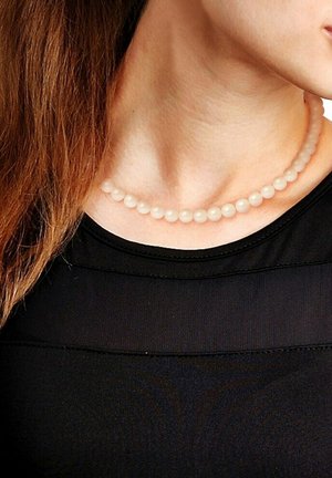 Collier de perles avec des perles rondes et lisses dans une couleur douce et pâle, reposant sur le cou, associé à un haut noir avec un tissu superposé.