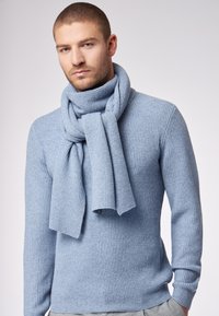 Uomo che indossa un pullover a coste di un blu chiaro e una sciarpa abbinata, davanti a uno sfondo chiaro e semplice, mentre guarda verso la fotocamera.