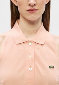Femme portant un polo sans manches de couleur pêche avec trois boutons et un petit logo de crocodile vert sur la poitrine.