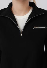 Personne portant un sweat-shirt noir à fermeture éclair partielle sur un t-shirt blanc côtelé à col rond, fermeture éclair partiellement ouverte, montrant le col et la poitrine.