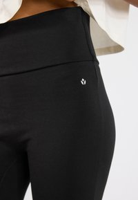 Colanți negri cu talie înaltă, din material elastic, având un detaliu mic cu logo alb pe şold și o textură netedă.
