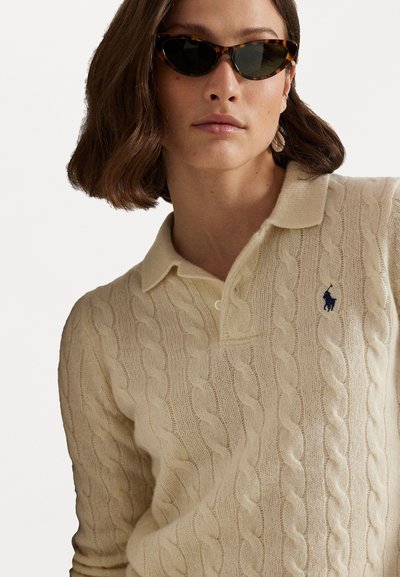 Crème kabelgebreide polo trui met een kraag en een klein blauw logo op de linkerborst, gecombineerd met schildpadbril in cat-eye stijl.