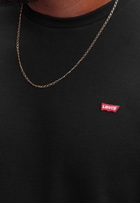T-shirt in cotone nero con un'etichetta rossa Levi's sul petto, scollatura rotonda e una collana d'oro visibile sopra.