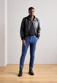Chaqueta bomber de cuero negro, camiseta negra de cuello redondo, pantalones ajustados azules y botas negras hasta el tobillo, todo ello sobre un fondo neutro.