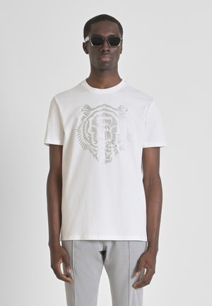 White cotton t-shirt featuring a raised grey lion graphic. Crew neckline, short sleeves, paired with light grey tailored trousers.