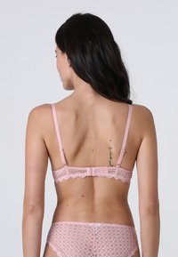 Donna vista di spalle che indossa un reggiseno di pizzo rosa e un perizoma di pizzo abbinato, con lunghi capelli scuri e un piccolo tatuaggio sulla parte superiore della schiena.