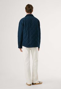 Chaqueta azul marino acolchada con cuello, mangas largas y puños abotonados, combinada con pantalones blancos y zapatos bajos, de pie frente a un fondo claro.