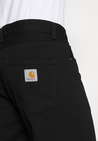 Pantaloni in denim nero con una tasca posteriore dotata di un'etichetta bianca che mostra un logo giallo. Il tessuto ha una texture liscia con dettagli di cucitura discreti.