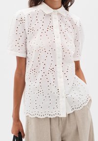 Chemisier blanc à manches courtes avec broderie ajourée, ourlet festonné, porté avec un pantalon beige, personne tenant une poignée de sac noire.