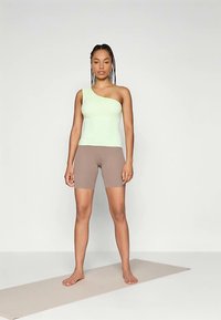 Top verde chiaro monospalla abbinato a pantaloncini da biker marroni. Tessuto liscio, design aderente. Modella in piedi a piedi nudi su un tappetino beige.