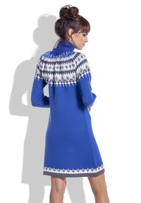 Kobieta z brązowymi włosami spiętymi w kok, ubrana w niebieską dzianinową sukienkę z białym i szarym wzorem na karczku i dole, stoi bokiem z rękami na biodrach.