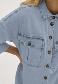 Chemise en denim bleu clair avec deux poches poitrine et des boutons marron, à coupe décontractée et avec des détails de coutures visibles.