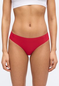 Γυναίκα που φοράει κόκκινο αόρατο εσώρουχο με χαμηλή μέση και λευκό κομψό τοπ, εμφανιζόμενη από το ύψος της μέσης έως τους μηρούς.