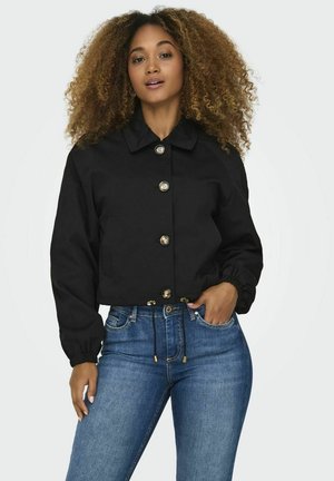 Black cropped jacket with a pointed collar, featuring gold buttons and elastic cuffs, worn over blue jeans. Smooth texture and relaxed fit.
