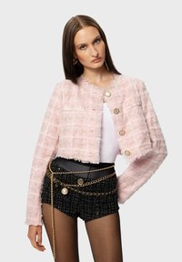Pink tweed cropped jacket featuring a textured grid pattern, frayed edges, and gold button closures. Worn with a black chain-accented belt.