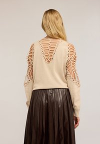 Donna con lunghi capelli biondi, che indossa un maglione beige con dettagli in crochet aperti sulla schiena e sulle maniche, abbinato a una gonna plissettata marrone scuro.