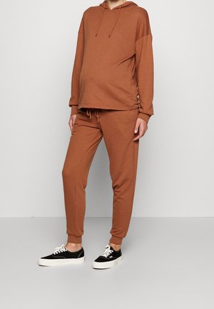 Osoba ubrana w rdzawej barwy bluzę z kapturem i dopasowane spodnie dresowe oraz czarno-białe sneakersy, stojąca na tle jednolitego, jasnoszarego tła.