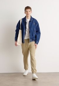 Jeansjakke med elastiske mansjetter og hem, iført over en hvit t-skjorte og beige joggebukser; bærer en kanvas tote bag; hvite sneakers.