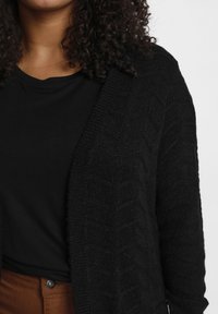 Schwarze Strickjacke mit strukturiertem Zickzackmuster, kombiniert mit einem schwarzen T-Shirt. Das Outfit wirkt warm und lässig.