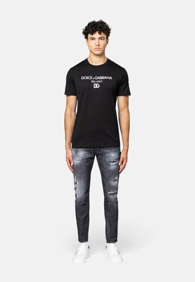 Zwarte katoenen t-shirt met witte merktekst, gecombineerd met versleten grijze spijkerbroek en witte sneakers. Model staat voor een neutrale achtergrond.