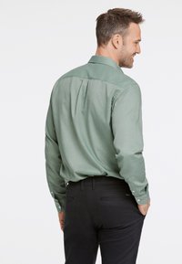 Camicia a maniche lunghe in morbido tessuto verde chiaro con una texture liscia, dotata di colletto classico e polsini abbottonati, abbinata a pantaloni neri scuri.