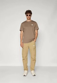 Muž v hnědém tričku s krátkým rukávem a béžových cargo kalhotách, v bílých teniskách a béžových slunečních brýlích, stojící na jednobarevném pozadí.