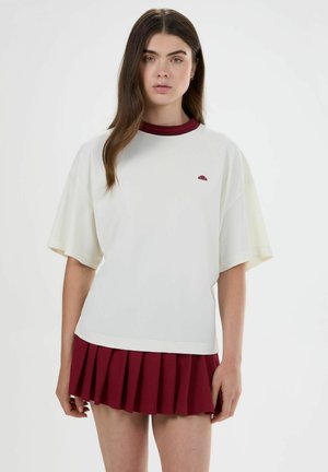 Joven mujer con cabello largo y castaño lleva una camiseta blanca holgada con un pequeño logo y una falda roja plisada, de pie frente a un fondo liso.