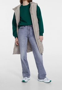 Béžová vycpávaná vesta s kapucí vrstvená přes zelenou mikinu, doplněná světle modrými džíny s širokými nohavicemi a bílými teniskami se zelenými prvky.