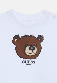 T-shirt en coton blanc avec un graphique de ours en cartoon marron et le nom de la marque "GUESS" avec "EST. 1981" en dessous.