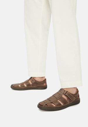 Persona con pantalones blancos y sandalias de cuero marrón cerradas con tiras de velcro, de pie sobre un fondo blanco.