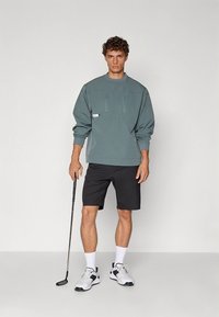 Světlé zelené mikina se dvěma kapsami na zip, v kombinaci s černými šortkami. Model drží golfovou hůl, má na sobě bílé ponožky a tenisky.