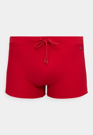 Piros fürdőnadrág rugalmas anyagból, elöl megkötővel, testhez simuló szabással, és egy kis logóval az oldalon.