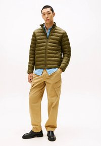 Olive green puffer jacket with horizontal quilting, zip closure, and stand collar. Worn over a light blue shirt and khaki cargo pants.