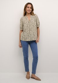 Blusa a maniche corte in colore crema con motivo marmorizzato marrone, con scollatura a V, abbinata a jeans skinny blu e ballerine beige.