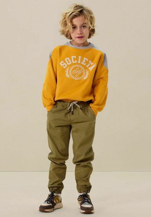 Sweatshirt jaune avec impression "SOCIETÀ", accents d'épaule gris, pantalon élastique vert olive à poignets resserrés et baskets multicolores.