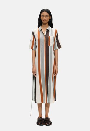 Kobieta stojąca w sukience do kolan z pionowymi paskami w kolorze białym, pomarańczowym, brązowym i szarym, zestawiona z ciemnymi, frędzlowanymi sandałami z odkrytymi palcami.