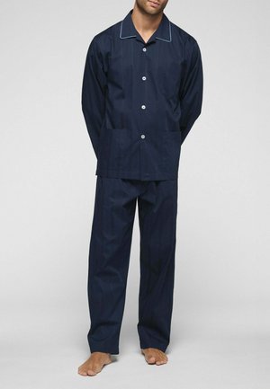Uomo in piedi a piedi nudi che indossa una maglia da pigiama blu navy a maniche lunghe con bottoni e pantaloni larghi abbinati su sfondo semplice.