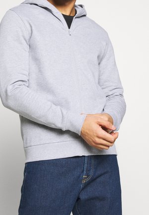 Mężczyzna ubrany w jasnoszarą bluzę z zamkiem na suwak noszoną na czarnej koszulce, w ciemnoniebieskich dżinsach, z rękami złączonymi przed sobą.