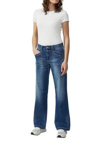 Frau in einem weißen kurzärmligen Oberteil, hoch taillierten blauen weit geschnittenen Jeans mit aufgesetzten vorderen Taschen und weißen Sneakers, die mit den Händen in den Taschen steht.