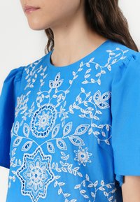 Blue cotton shirt with puffed sleeves, featuring intricate white floral embroidery and symmetrical patterns on the front and shoulders.