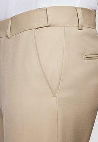 Beżowe spodnie o gładkiej teksturze, z prostym krojem, ukośnymi kieszeniami z przodu i subtelnym diagonalnym wzorem w tkaninie.