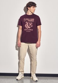 Bordová bavlněná tričko s korálovým grafickým motivem v světlé růžové, doplněné béžovými kalhotami a krémovými teniskami, na neutrálním pozadí.